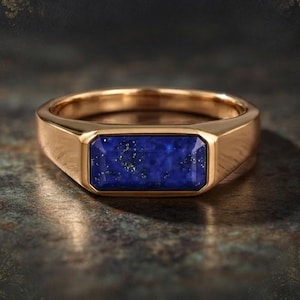 May include: A gold ring featuring a rectangular, deep blue lapis lazuli gemstone set horizontally. The stone displays natural gold-colored flecks across its surface, and the band has a smooth, polished finish with a flat, wide top section.