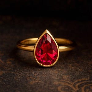 Op de afbeelding: Een gouden ring met een peervormige, gefacetteerde, helderrode edelsteen. De ring heeft een eenvoudige, gepolijste gouden band en een bezel setting die de levendige kleur van de steen benadrukt. De ring wordt getoond op een donker, patroon oppervlak.
