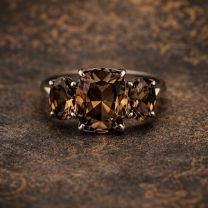 Op de afbeelding: Een ring met drie bruine edelstenen. De centrale steen is rechthoekig, met twee ovale stenen aan weerszijden. De ring is geplaatst tegen een donkere, patroonachtergrond.