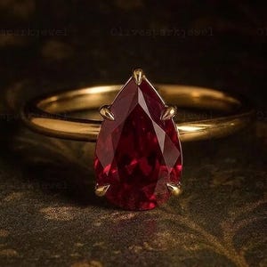 May include: A gold ring featuring a pear-shaped, deep red gemstone set in a six-prong mounting. The band is smooth and polished, showcasing a classic design with a faceted stone that reflects light, centred on the setting with a pointed top.