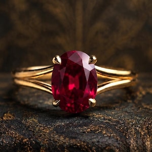 Könnte beinhalten: Ein Goldring mit einem zentralen, oval geschliffenen, tiefroten Edelstein, der von vier Krappen gehalten wird. Die Ringschiene besteht aus poliertem Gelbgold und teilt sich zur Fassung hin in zwei Abschnitte, was für ein elegantes Design sorgt.