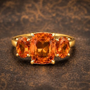 May include: A gold ring featuring a large, cushion-cut orange gemstone in the center, flanked by two smaller, oval-cut orange gemstones on either side. The stones are set in a polished gold band with four prongs securing the central stone.