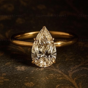 May include: A gold ring with a pear-shaped diamond. The diamond is the focal point, set in a classic prong setting. The band is a simple, polished gold. The ring is displayed on a dark, patterned surface.