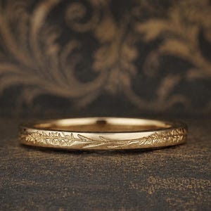 Könnte beinhalten: Ein goldfarbener Ring mit einem eingravierten floralen Muster. Der Ring ist vor einem dunklen, gemusterten Hintergrund platziert. Das Band zeigt ein durchgehendes Design aus Blättern und zarten Stielen.
