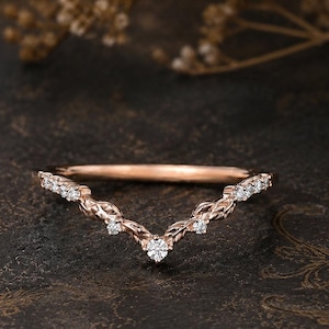 May include: A rose gold chevron ring with small, clear gemstones. The ring has a delicate, intertwined design and is set against a dark, textured background. The ring is a piece of jewelry.