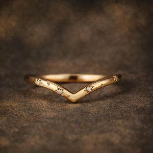 Könnte beinhalten: Ein goldfarbener, V-förmiger Ring mit kleinen, funkelnden Edelsteinen und sternförmigen Gravuren. Der Ring ist vor einem dunklen, strukturierten Hintergrund platziert, der sein zartes Design und seine Handwerkskunst hervorhebt. Dieser Ring ist ein Schmuckstück.
