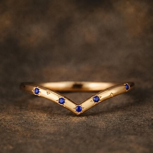 May include: A gold V-shaped ring featuring four small, round, deep blue stones set into the band. Each stone is surrounded by a starburst engraving, with additional small star engravings interspersed along the polished gold surface of the ring.