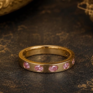 Può includere: Un anello a fascia in oro con una serie di pietre preziose rosa di forma ovale incastonate a filo lungo la circonferenza esterna. Il metallo ha una finitura lucida e le pietre sono disposte in un motivo ripetitivo lungo tutto l'anello.