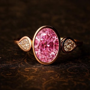 May include: Rose gold ring featuring a large, oval-cut pink gemstone as the centerpiece, flanked by two smaller, pear-shaped, clear gemstones. The ring's band and setting are a warm, metallic color, creating a striking contrast with the vibrant pink of the main stone.