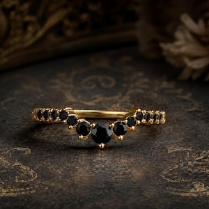 Könnte beinhalten: Ein goldener Ring mit einem geschwungenen, V-förmigen Design, besetzt mit einer Reihe runder, facettierter schwarzer Steine. Der mittlere Stein ist etwas größer als die umliegenden Steine, die symmetrisch entlang des Bogens angeordnet sind.