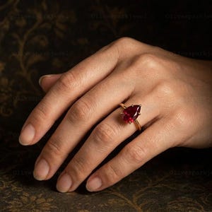 May include: A gold ring featuring a single pear-shaped, deep red gemstone set in a simple prong setting. The band is thin and polished, showcasing the faceted cut of the stone which reflects light across its surface. The design is clean and classic.