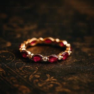 May include: A gold ring with a repeating pattern of oval-cut, deep red gemstones and small, round, clear gemstones. The ring is displayed on a dark, textured surface, enhancing the contrast and the ring's design.