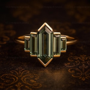 May include: A gold ring featuring a central green gemstone with a geometric design. The ring has a rectangular center stone flanked by smaller rectangular stones, all set in a gold band. The ring's design is Art Deco inspired.