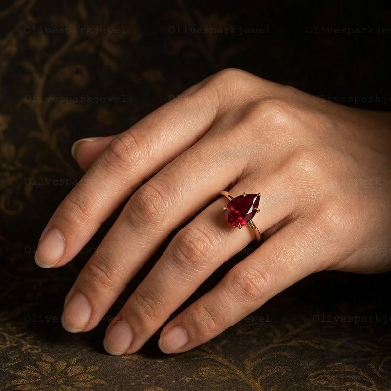 May include: A gold ring featuring a single pear-shaped, deep red gemstone set in a simple prong setting. The band is thin and polished, showcasing the faceted cut of the stone which reflects light across its surface. The design is clean and classic.