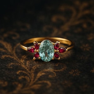 May include: A gold ring featuring an oval, light blue gemstone at the center, surrounded by six smaller, red gemstones in a floral arrangement. The ring's band is a simple, polished gold.