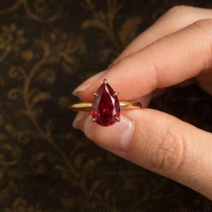 May include: A gold ring featuring a single pear-cut deep red gemstone held in a six-prong setting. The band is smooth and polished, showcasing a minimalist design that highlights the vibrant color and faceted surface of the central stone.