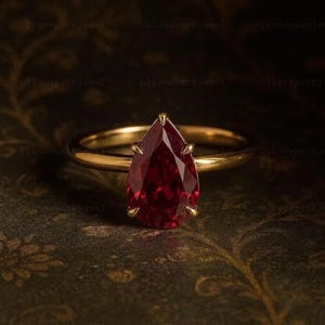 May include: A gold ring featuring a pear-shaped, deep red gemstone set in a six-prong mounting. The band is smooth and polished, showcasing a classic design with a faceted stone that reflects light, centered on the setting with a pointed top.