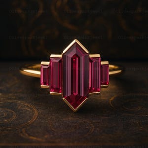 May include: A gold ring with a central, rectangular, deep red gemstone, with two smaller matching gemstones on either side. The ring has a geometric design and a polished gold band. The gemstones are a rich, vibrant colour.