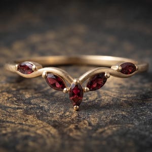 Op de afbeelding: Een gouden ring met een V-vormige band bezet met vijf dieprode granaat edelstenen in marquise-slijpvorm. Het metaal heeft een matte afwerking en de stenen zijn in een symmetrisch patroon gerangschikt langs het gebogen bovenste deel van de ring.
