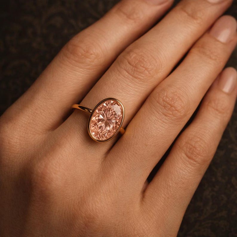 May include: A gold ring featuring a large, oval-cut, peach-colored gemstone. The ring's band is a thin, polished gold, and the gemstone is set in a bezel setting. The ring is displayed on a hand with well-manicured nails.