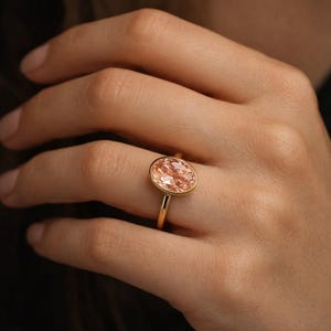 May include: A gold ring featuring a large, oval-cut peach-colored gemstone. The ring has a thin, polished gold band and a bezel setting that surrounds the gemstone. The ring is shown on a hand.