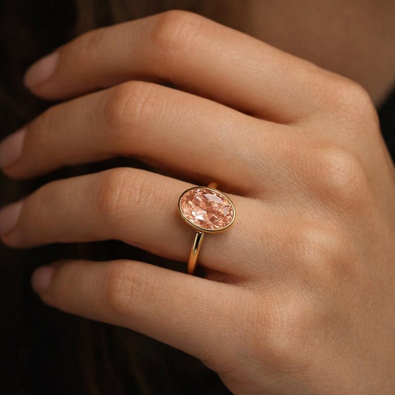 May include: A gold ring featuring a large, oval-cut peach-colored gemstone. The ring has a thin, polished gold band and a bezel setting that surrounds the gemstone. The ring is shown on a hand.