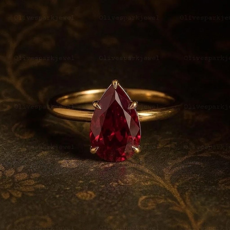 May include: A gold ring featuring a pear-shaped, deep red gemstone set in a six-prong mounting. The band is smooth and polished, showcasing a classic design with a faceted stone that reflects light, centered on the setting with a pointed top.