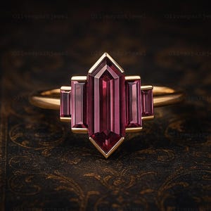 May include: A gold ring featuring a central rectangular, deep red gemstone flanked by two smaller rectangular gems on each side. The ring has a geometric design with a pointed bottom and a gold band.