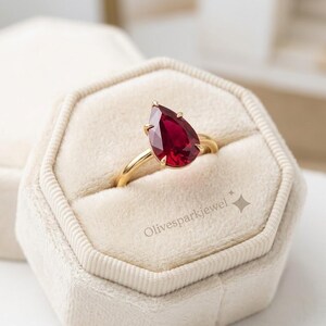 May include: A gold ring featuring a pear-shaped deep red gemstone set in a prong setting. The ring is displayed in a beige velvet hexagonal box. The text Olivesparkjewel is printed on the surface of the box near the ring.