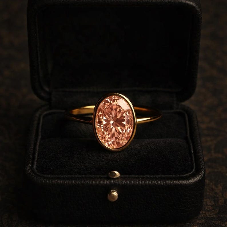 May include: A gold ring with an oval, peach-colored gemstone set in a bezel setting. The ring is displayed in a black jewelry box with a dark interior. The ring's band is thin and polished.