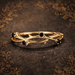 May include: A gold-colored ring with a branch-like design. The ring features four small, round, black gemstones. The band has a delicate, intertwined branch pattern with pointed details. The ring is set against a dark, patterned background.
