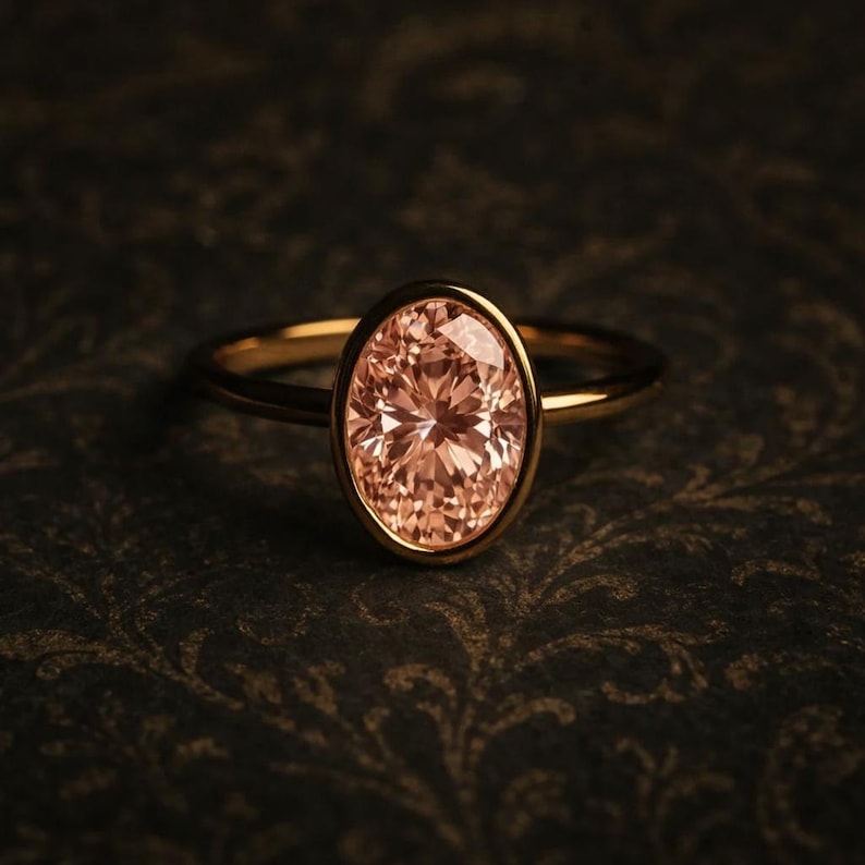 May include: A gold ring featuring an oval-cut, peach-colored gemstone. The gemstone is set in a bezel setting, and the band is a thin, polished gold. The ring is displayed against a dark, patterned background.