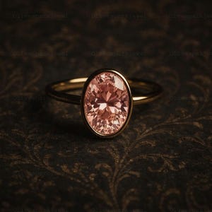 May include: A gold ring featuring an oval-cut, light pink gemstone. The gemstone is set in a bezel setting, and the band is a simple, thin design. The ring is displayed on a dark, patterned surface.
