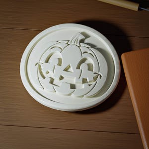 May include: A circular white cookie cutter featuring a raised, embossed design of a jack-o'-lantern pumpkin with a carved face, including triangular eyes, a triangular nose, and a wide, jagged mouth with square teeth, set within a round frame.