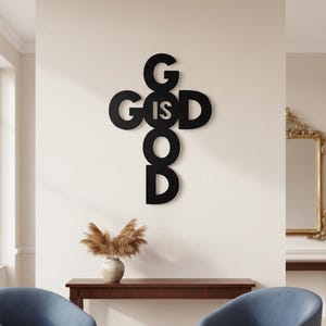 May include: Black metal wall art in the shape of a cross with the words "GOD IS GOOD" cut out. The art is mounted on a cream-colored wall above a wooden table. A vase with dried plants and two blue armchairs are in the foreground.