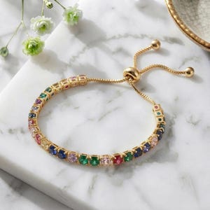 Puede incluir: Una pulsera tipo bolo ajustable en tono dorado con una hilera continua de piedras redondas multicolor en tonos azul, verde, rosa y amarillo. La pulsera incluye un mecanismo deslizante con dos cadenas doradas colgantes en el extremo.