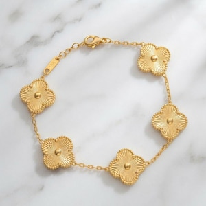 May include: A gold-toned bracelet featuring five clover-shaped charms with a radial ribbed texture and a central raised dot. The chain links connect each charm, and a small rectangular tag stamped with 18K is attached near the lobster clasp.