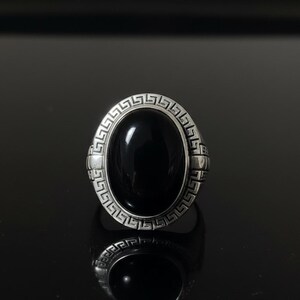May include: A silver ring with a large, oval, black stone. The ring has a detailed, geometric design around the stone and on the sides. The ring is displayed on a reflective black surface.