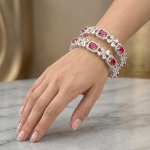 May include: Two silver-toned bracelets featuring rectangular, deep red stones set in a frame of small, clear, sparkling stones. The design alternates between the red stone settings and four-petaled flower shapes made of clear, faceted stones.