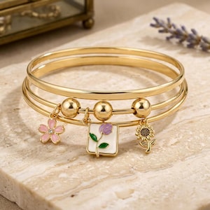 Può includere: Un bracciale rigido color oro composto da tre sottili cerchi sovrapposti. Il bracciale è decorato con tre ciondoli pendenti: un fiore in smalto rosa, un ciondolo quadrato in smalto bianco con un tulipano viola e un ciondolo a forma di girasole.
