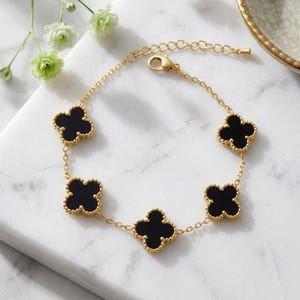 May include: A gold-tone chain bracelet featuring five evenly spaced black clover-shaped charms with beaded edges. The bracelet includes a delicate link chain and a lobster claw clasp, creating a symmetrical design with a polished finish.