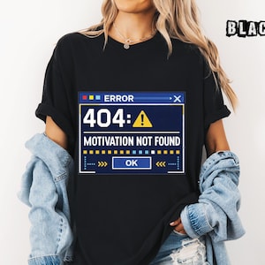 May include: Black t-shirt featuring a computer error message. The message reads "ERROR 404: MOTIVATION NOT FOUND" in a blue and white window design with an exclamation point symbol. The word "OK" is also visible.