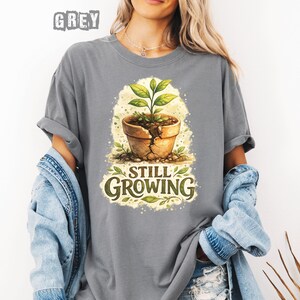 May include: A grey t-shirt with a watercolor illustration of a potted plant with the words "Still Growing". The plant pot is cracked, and the plant has green leaves. The word "GREY" is printed at the top.