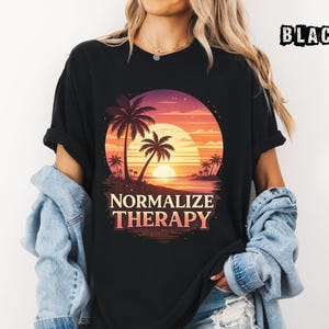 May include: Black t-shirt with a sunset graphic, palm trees, and the text "NORMALIZE THERAPY". The design uses orange, pink, and purple hues. The shirt is worn with a denim jacket. The word "BLACK" is printed on the top right.