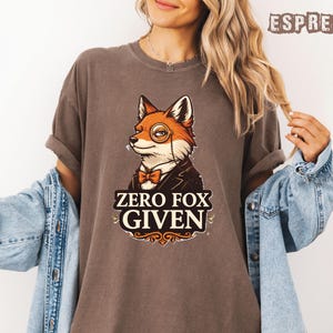 May include: Espresso-colored t-shirt featuring a cartoon fox wearing a monocle, bow tie, and suit jacket. The text "ZERO FOX GIVEN" is displayed below the fox. The shirt is a relaxed fit.