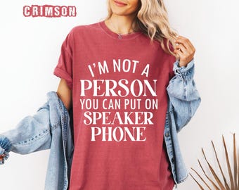 May include: Crimson red t-shirt with the text "I'M NOT A PERSON YOU CAN PUT ON SPEAKER PHONE" in white block letters. The shirt is a solid color and has short sleeves. A light blue denim jacket is draped over the shoulders.