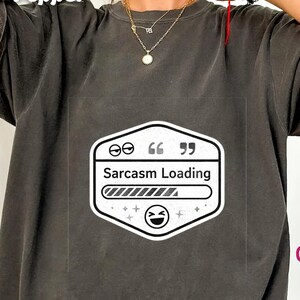 May include: Dark gray t-shirt with a white and black graphic that reads "Sarcasm Loading." The graphic features a loading bar, quotation marks, and a smiling face. The shirt is a casual style.