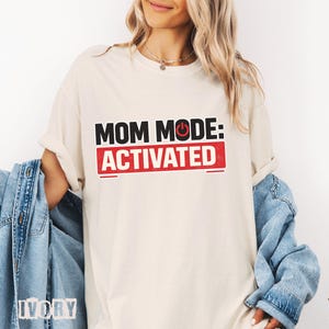 May include: Ivory-colored t-shirt with the text "MOM MODE: ACTIVATED" in black and red. The shirt has a relaxed fit and rolled-up sleeves. A denim jacket is draped over the arm.
