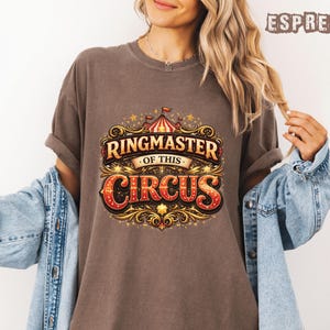 May include: Espresso-colored t-shirt with a vintage circus design. The graphic reads "Ringmaster of this Circus" in a decorative font with gold and red accents. The shirt has rolled-up sleeves and a relaxed fit.