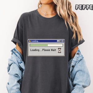 May include: Dark grey t-shirt with a retro computer loading screen graphic. The graphic shows a progress bar and the text "Loading... Please Wait". The shirt has short sleeves and a relaxed fit.